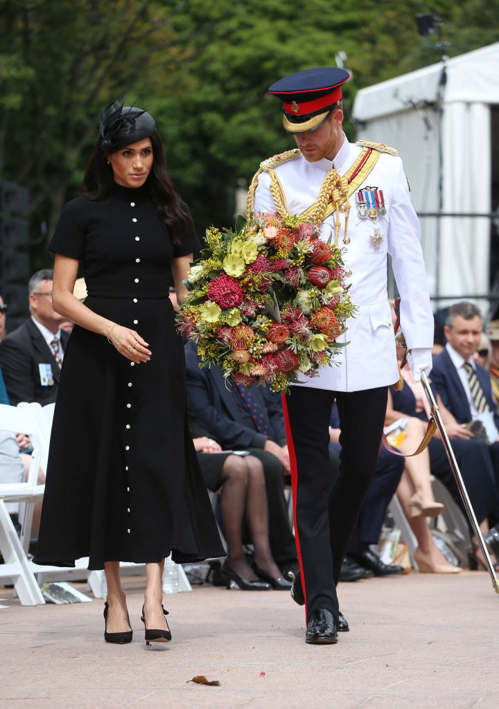 Harry wears a military uniform while carrying a bouquet of flowers and Meghan wears a dress with heels and a hat