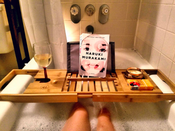 A wooden tray placed over a bubble bath holding a glass of wine, a book and a candle