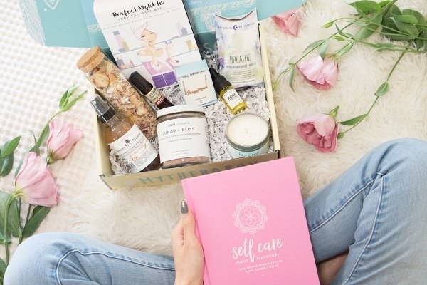 a person holding a self-care wellness book next to the box which is filled with a candle, essential oils, and other products