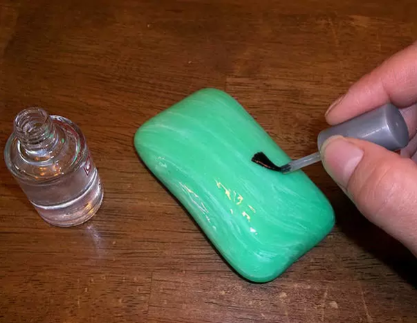 Hand painting a layer of clear polish on a green bar of soap, next to an open polish bottle