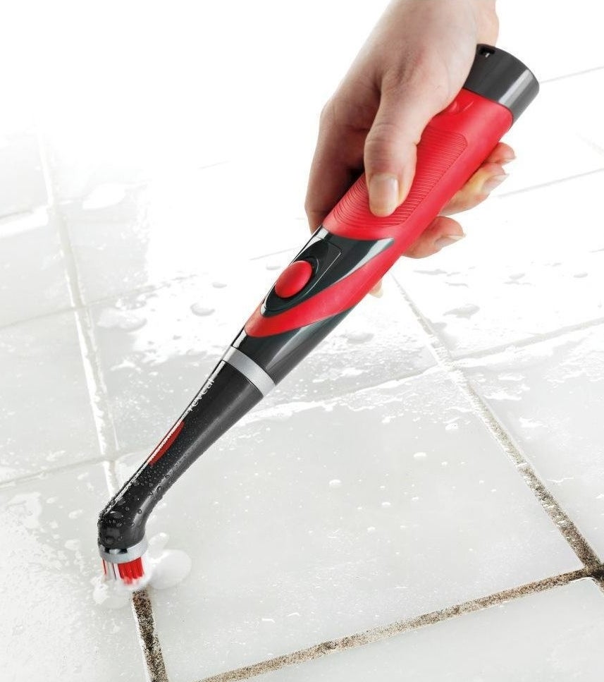 Hand holding the brush, which looks like an oversized electric toothbrush, while it scrubs dirty grout until it&#x27;s white