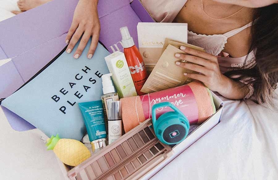a person holding the subscription box which has a Bluetooth speaker, towel, pillow, makeup palette, and other products inside it