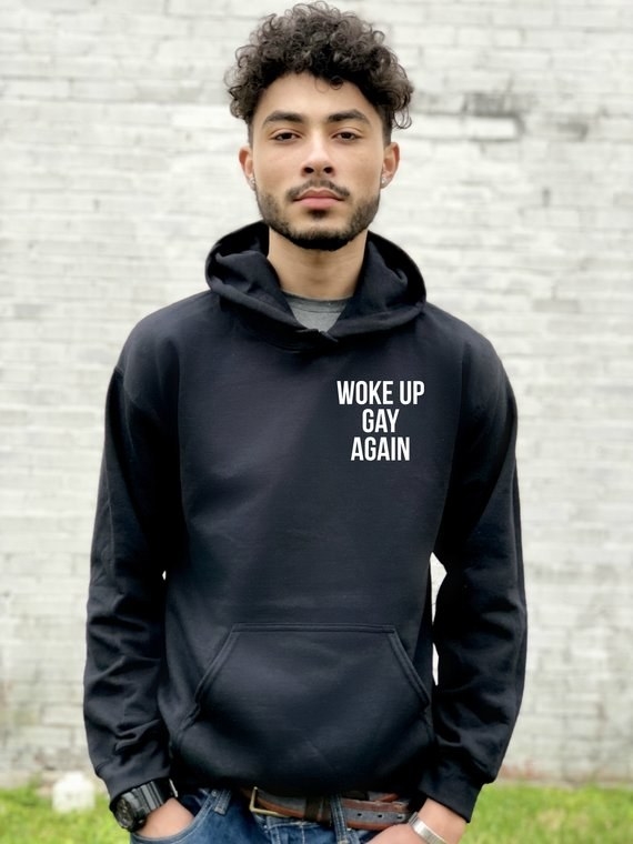 a model wearing a black hoodie that reads &quot;woke up gay again&quot; in white text