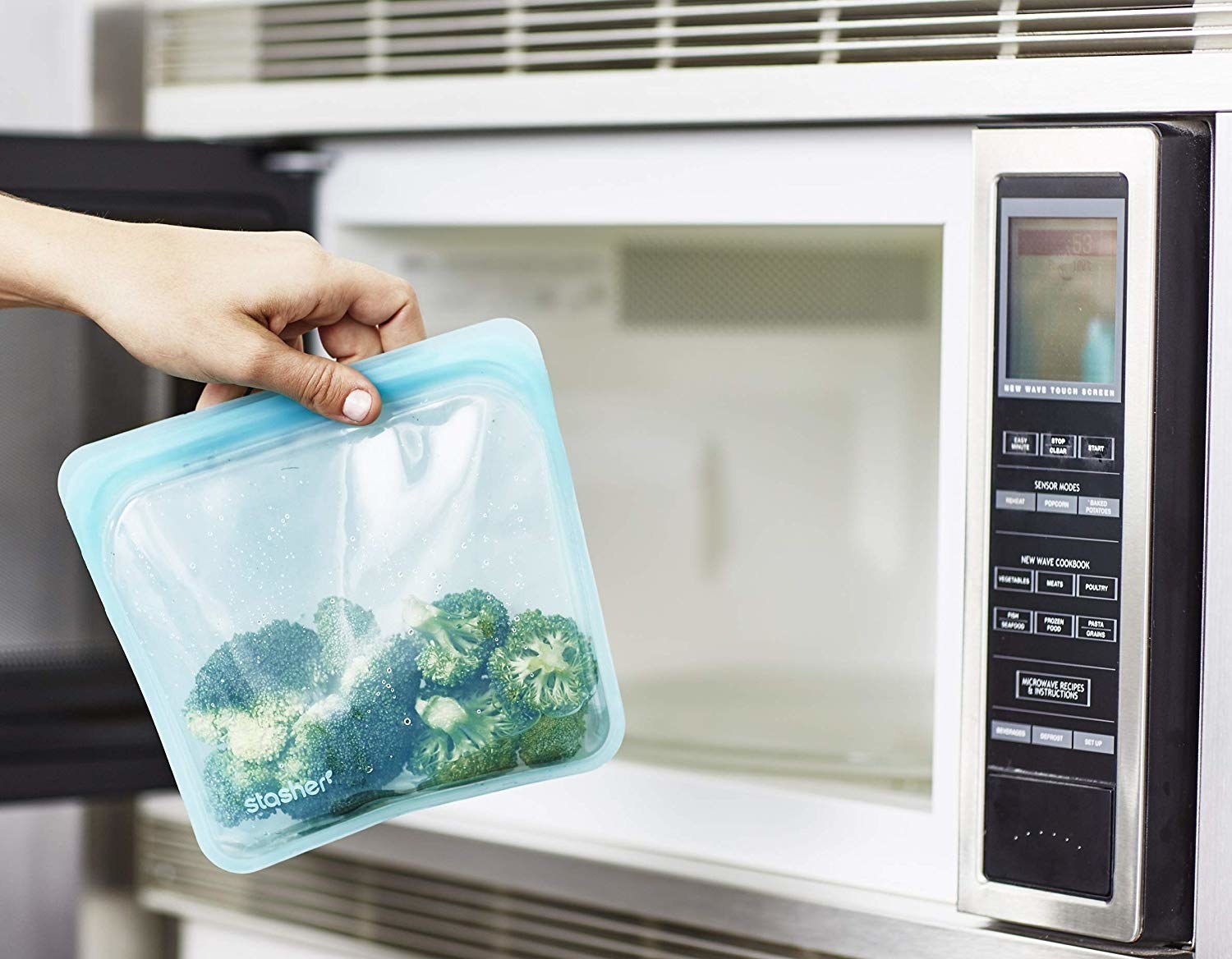 model&#x27;s hand puts clear blue stasher bag full of broccoli into microwave