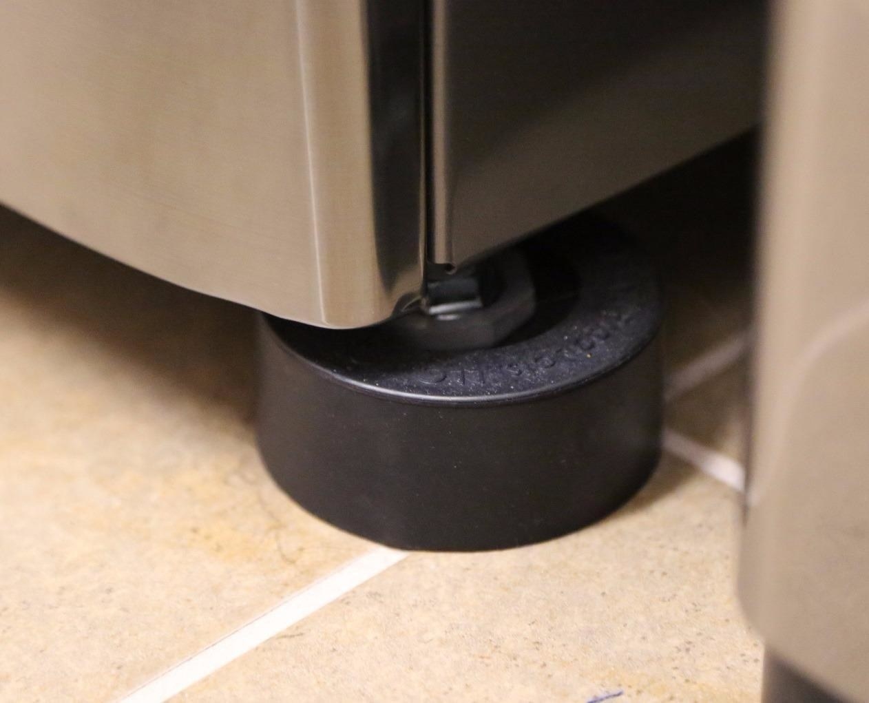 reviewer pic of black rubber hockey puck-like pad underneath the corner of a washing machine on a tile floor