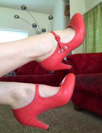 A reviewer wearing the shoes in red. They have a rounded toe, a high heel, a scallop cut-out detail on the sides, and a rounded red strap closure. 