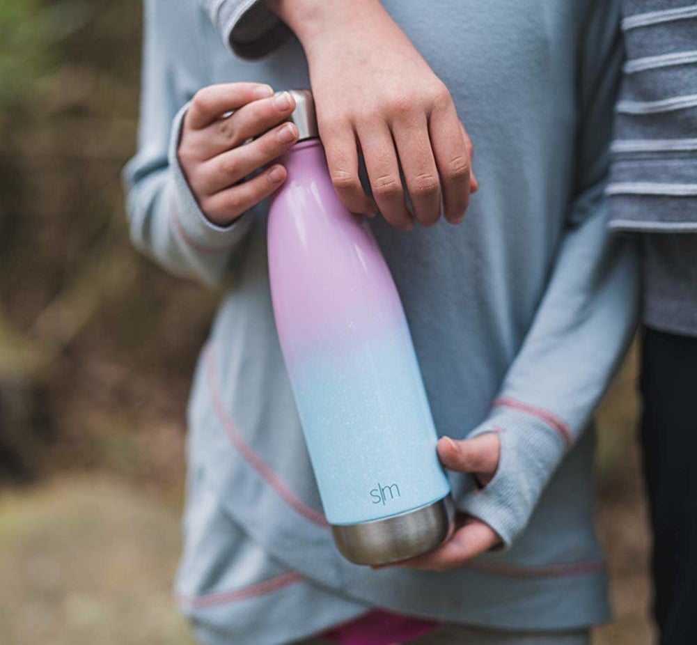 hands holding thin, long stainess steel bottle with a light pink to light blue gradient; it tapers at the top to a narrower opening with lid