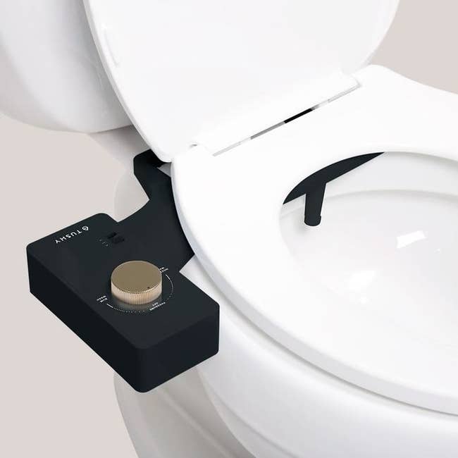 Black bidet attachment hooked to toilet seat. It has a gold knob to adjust pressure. 
