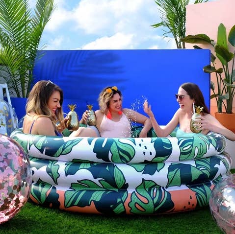 a group of three adults drinking out of pineapple cups and splashing into a mini blow up pool with a leafy design all over it