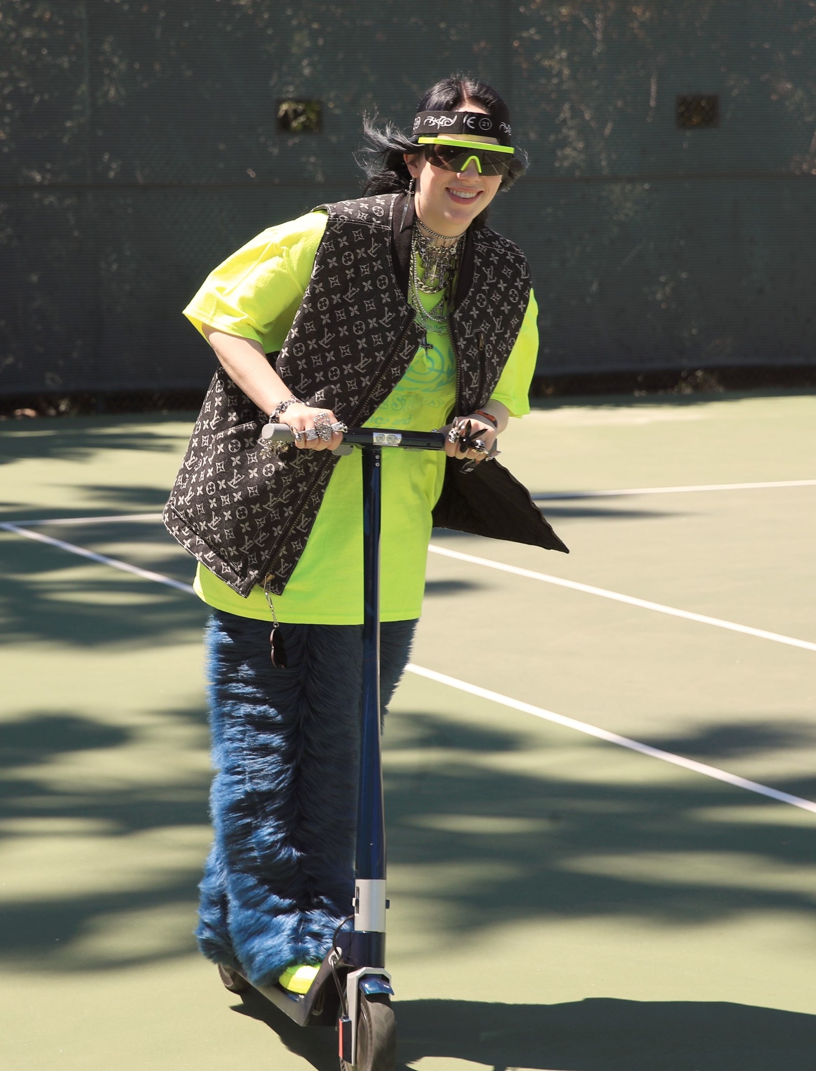 Billie com uma calça azul felpuda, camiseta verde neon e colete da Louis Vuitton andando de patinete