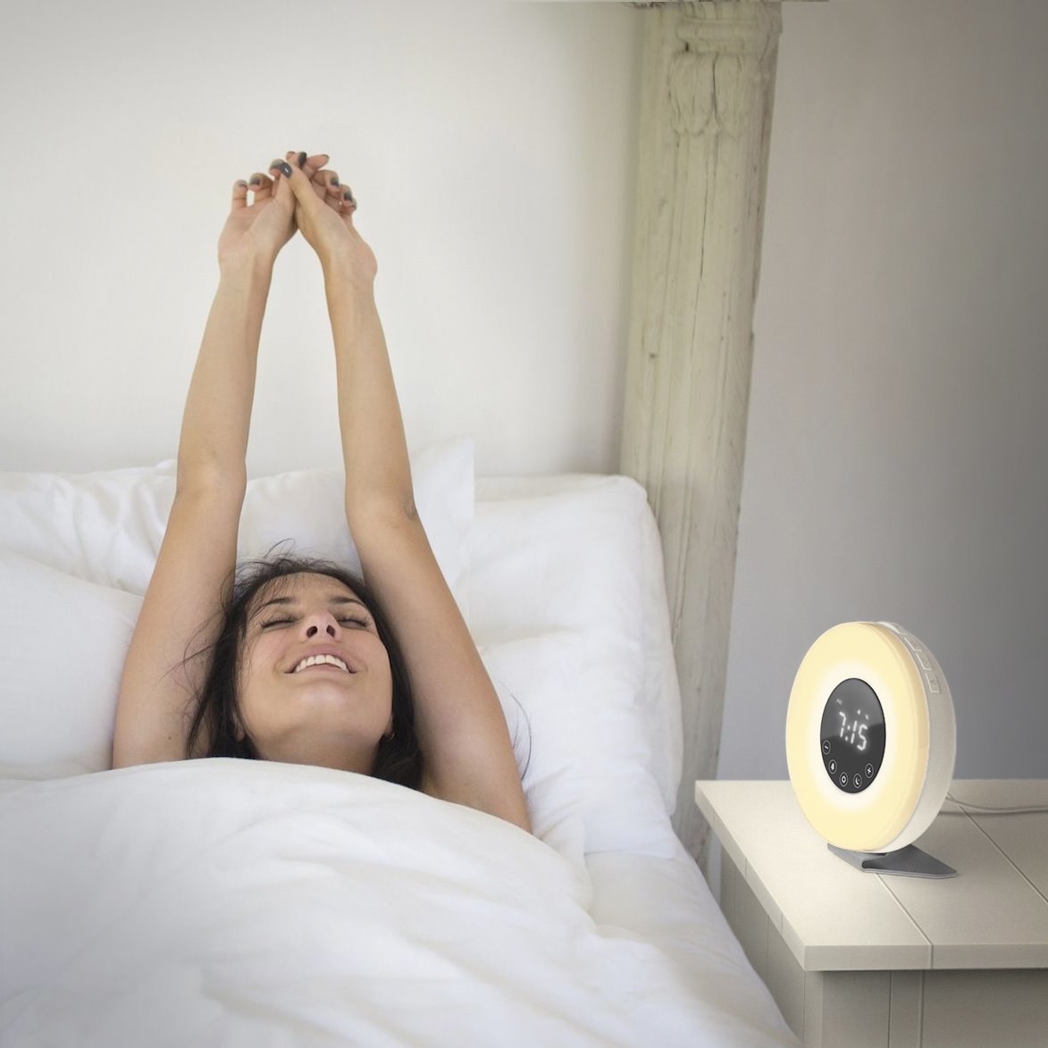 The circular alarm clock with an outer ring of light and inner black circle with the time on it sitting on a bedside table with a model in bed next to it stretching their arms