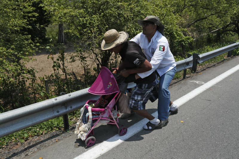 A Central American migrant pushing a child in a baby carriage is detained by a Mexican immigration agent from behind.