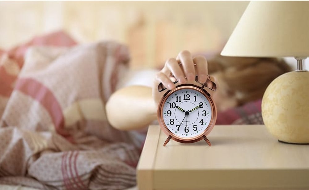 copper color alarm clock on night stand with person in bed reaching over to turn it off