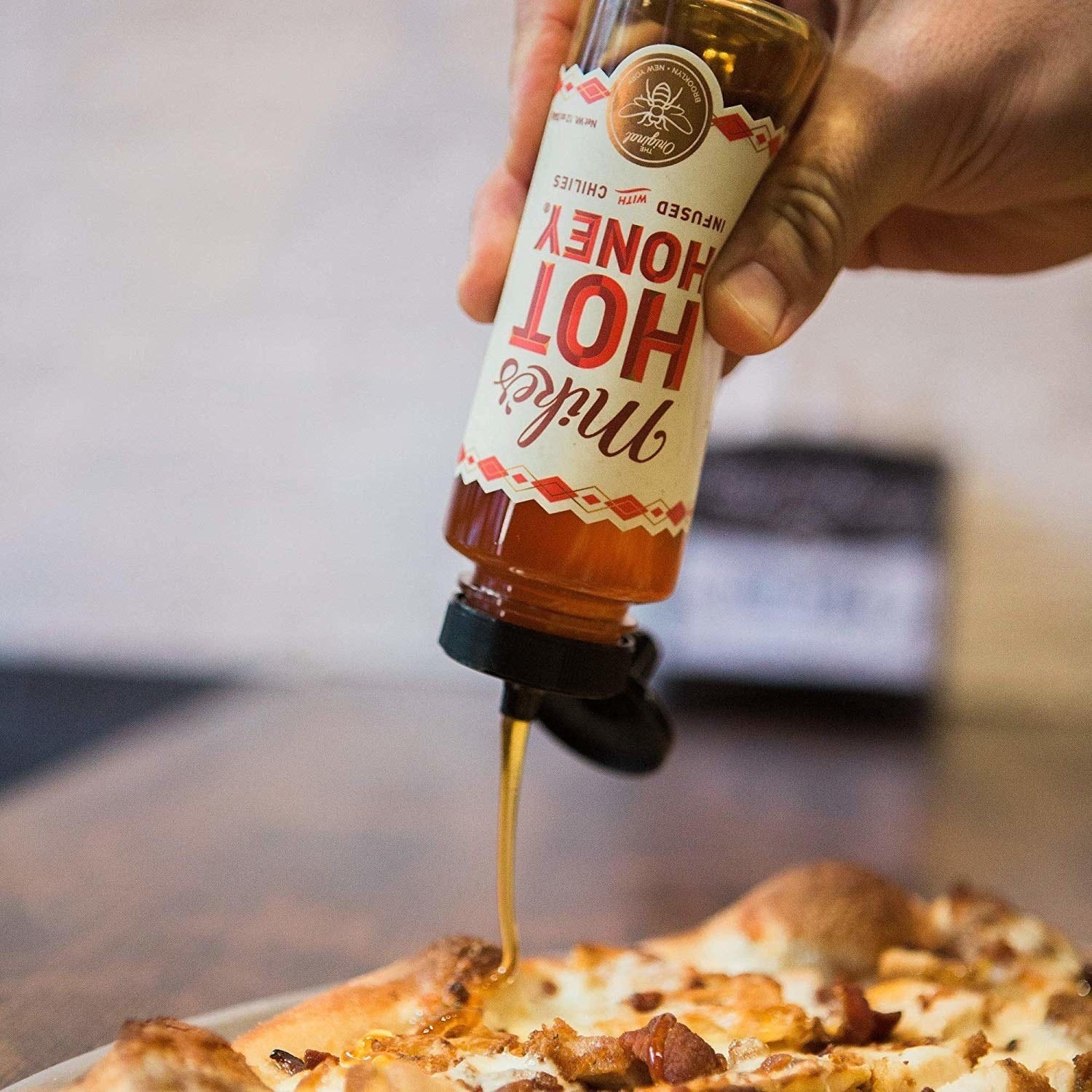 A hand holding the 12 ounce (340 gram) bottle upside down, drizzling the honey over pizza. The bottle has the name of the honey and says &quot;infused with chiles&quot;