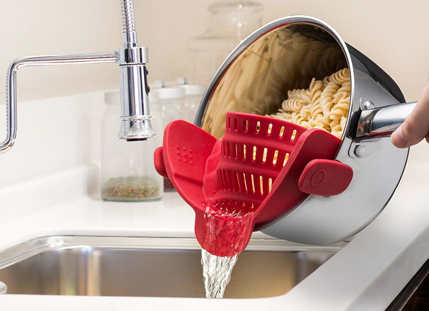 One hand pouring water out of a pot of pasta using the red strainer