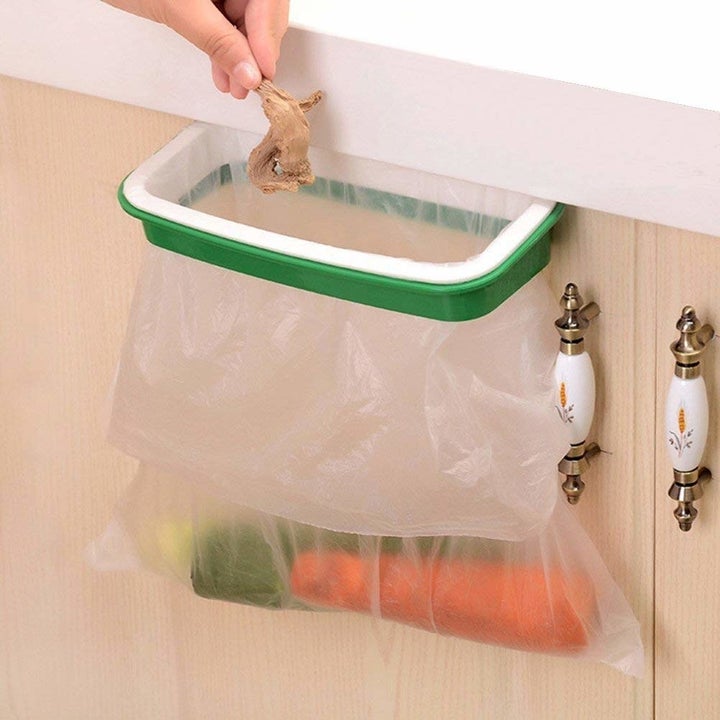 The holder clipped to a cabinet door, with a plastic bag inside and a hand putting food scraps inside it