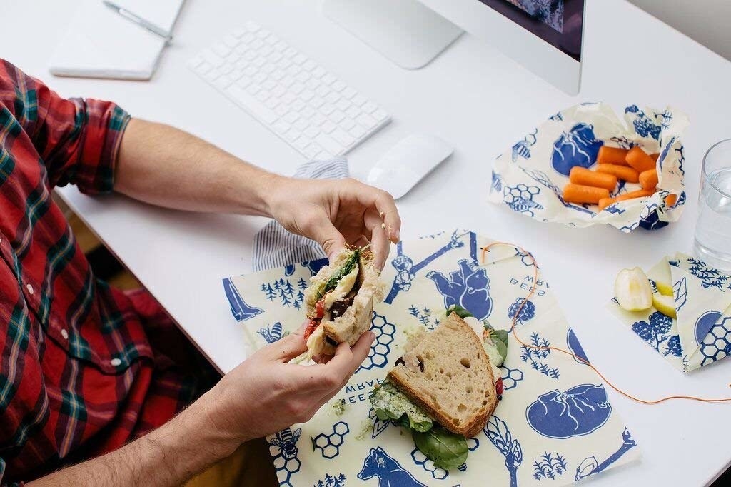 A lifestyle shot of three pieces of wax wrap being used to store a sandwich, carrots, and apple slices 