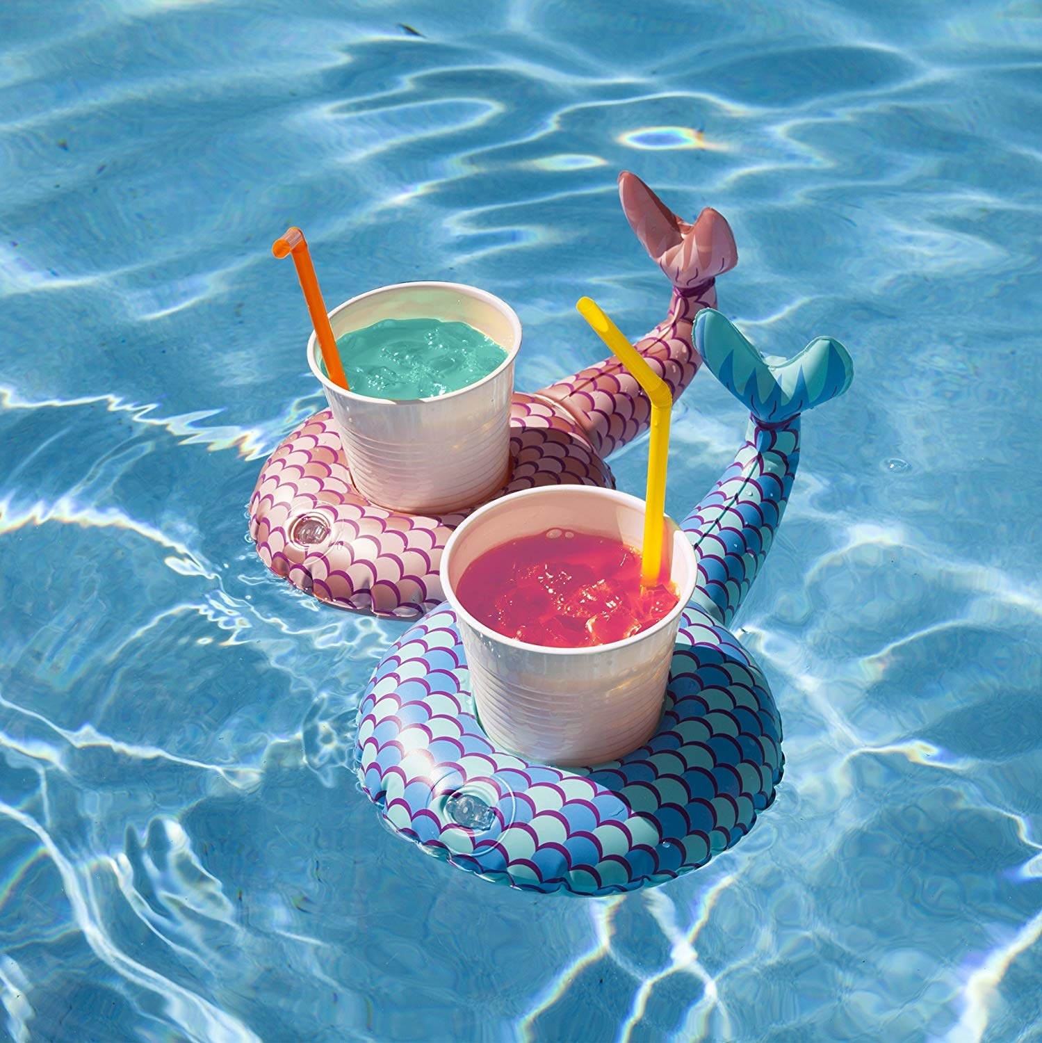The two drink floats in pink and blue printed with mermaid scales and with tails coming out, floating in a pool holding drinks