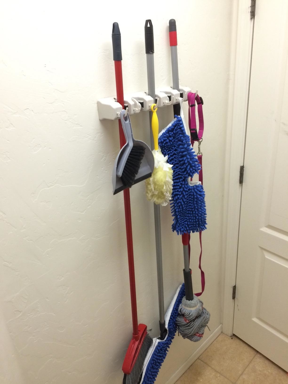 The white rack mounted behind a door with a broom and two styles of mop; plus a Swiffer, a mini broom and dustpan, and the dog&#x27;s leash
