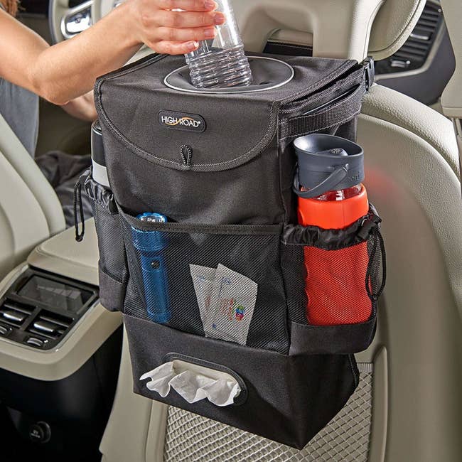 the car storage bag mounted to the back of the passenger seat with a hand putting a bottle into the top compartment, side pockets with water bottles inside, and we wipes peeking out of a dispenser along the bottom 