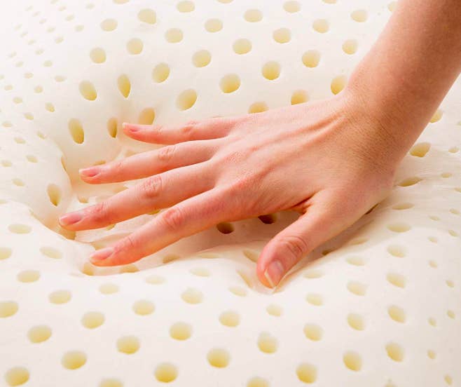 A model&#x27;s hand pressing against the pillow, creating a deep shape where their hand was to show the forming memory foam. The pillow has holes throughout it for structure and circulation. 