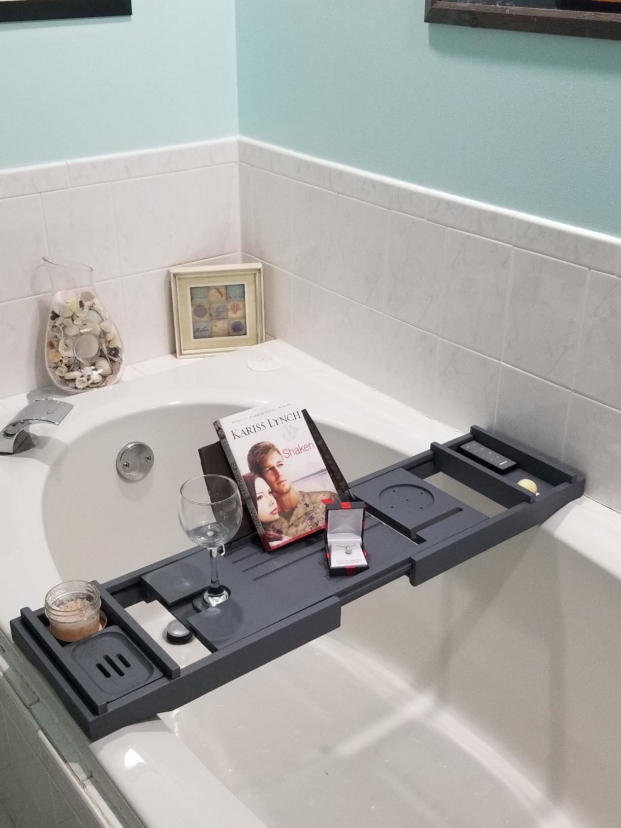 A reviewer&#x27;s bath caddy in grey with a candle wine glass and book on top in the set slots