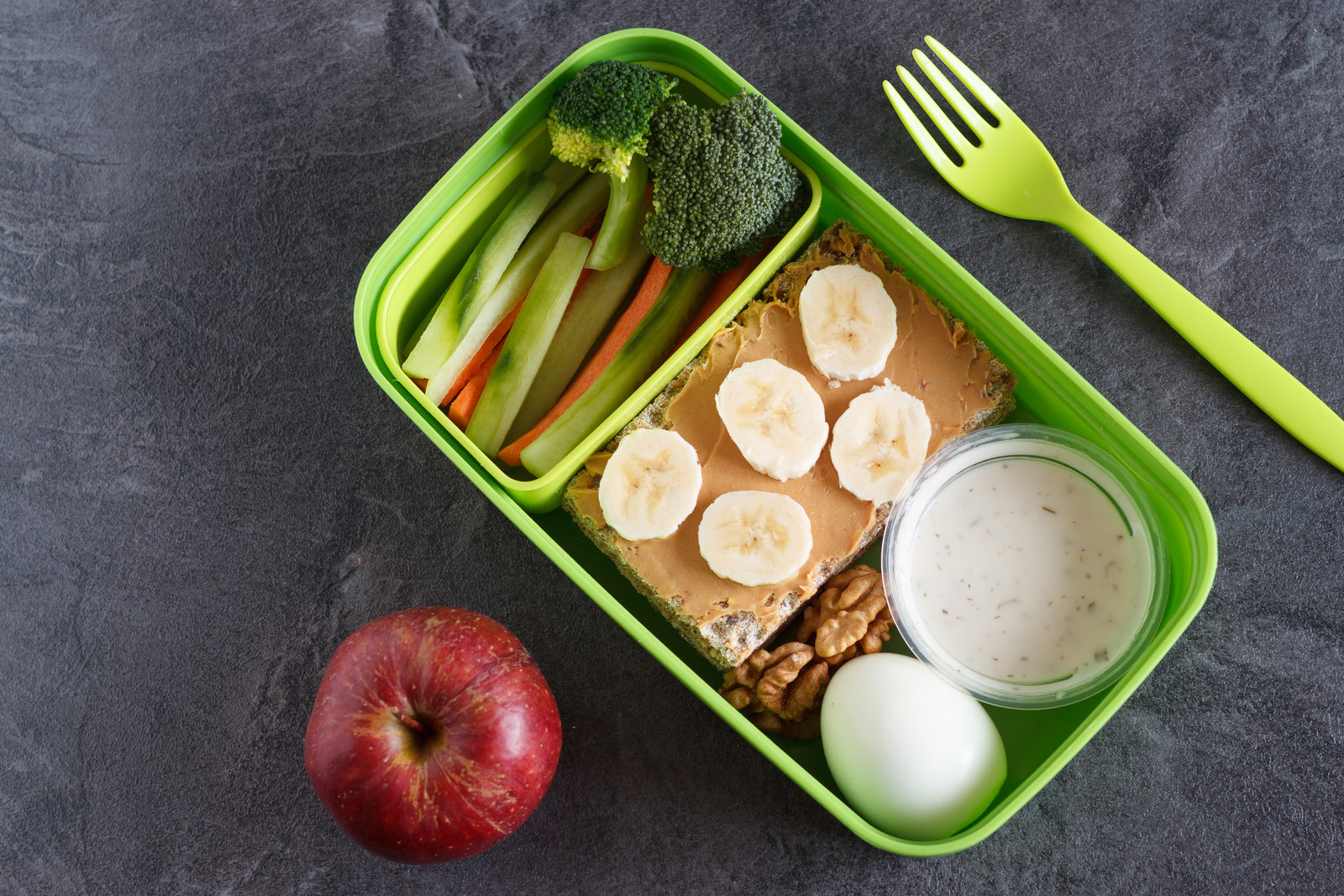 An open lunchbox containing a peanut butter sandwich, hard-boiled egg, and raw veggies with an apple on the side