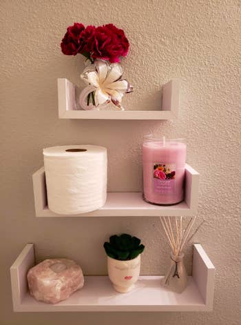 Closeup of three shelves vertically placed on a wall with smallest on top with assortest bathroom items on them, including toilet paper, a candle, etc. 