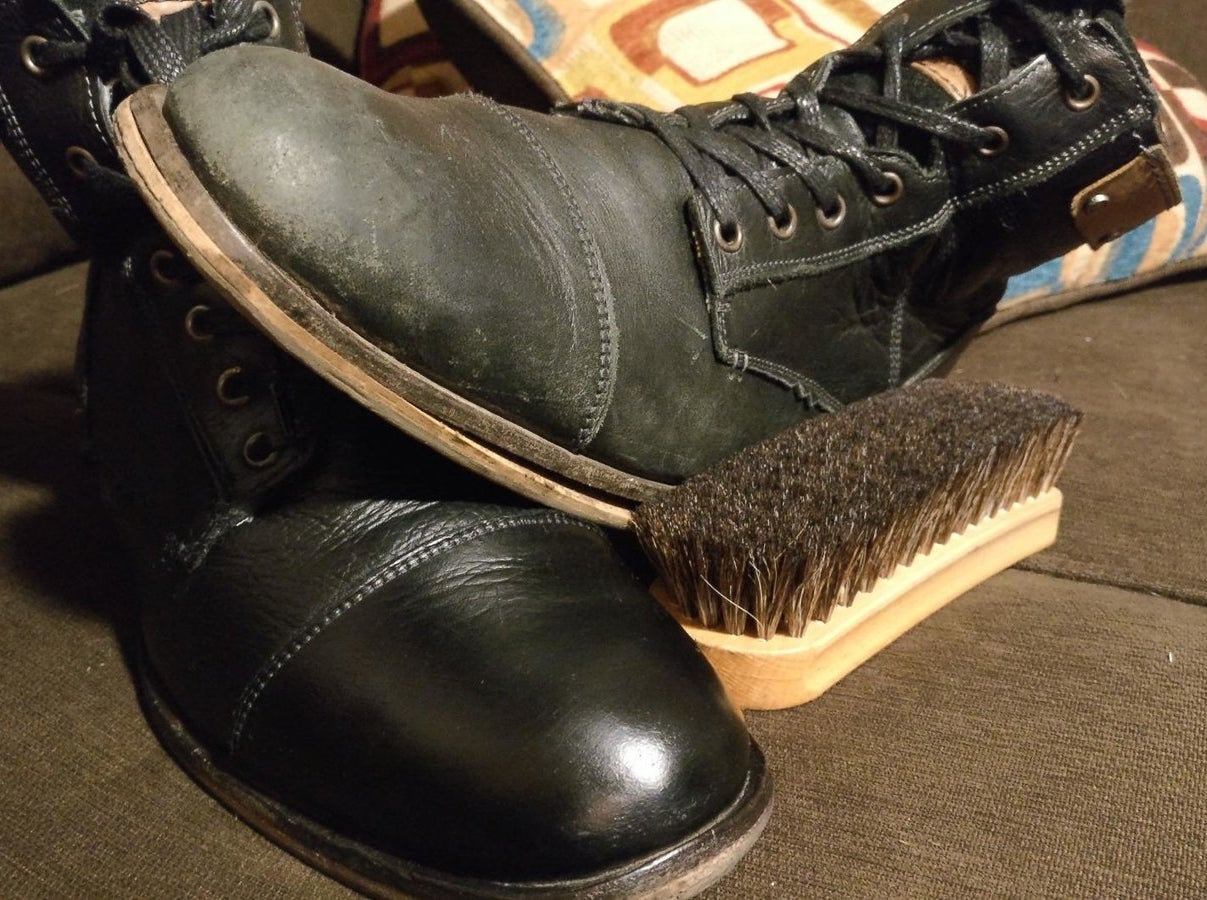 a scuffed black boot sitting on top of a shiny black boot to show contrast with a scrub brush next to them