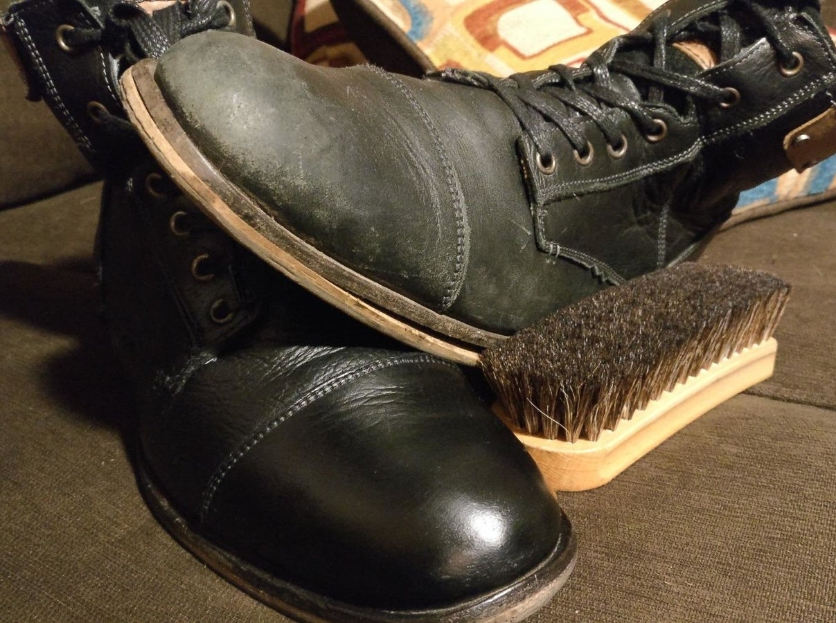 a reviewer&#x27;s scuffed black boot sitting on top of a shiny black boot to show contrast with a scrub brush next to them