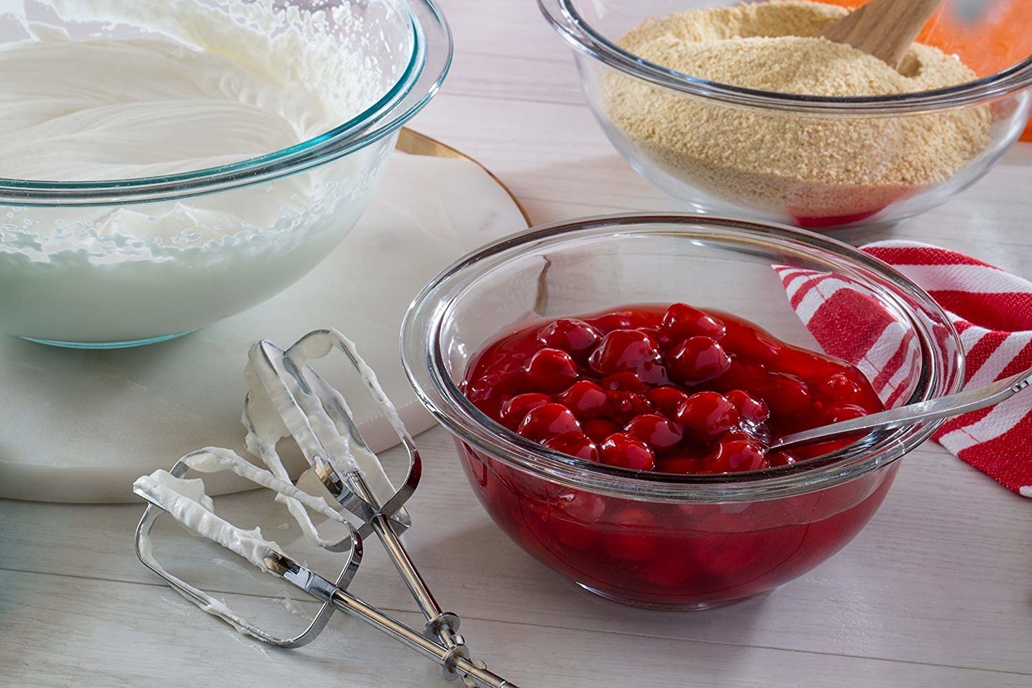 Bowls with whipped cream, cherries, and breadcrumbs, and a hand mixer on a table, ingredients for a dessert recipe