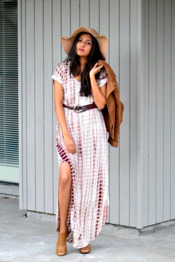 A reviewer wearing the brown and white dress with a belt and floppy hat, and sticking their leg through the high slit