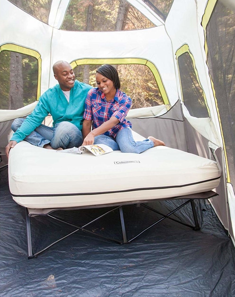 two models sitting on a queen-size inflatable mattress on a cot inside a tall tent