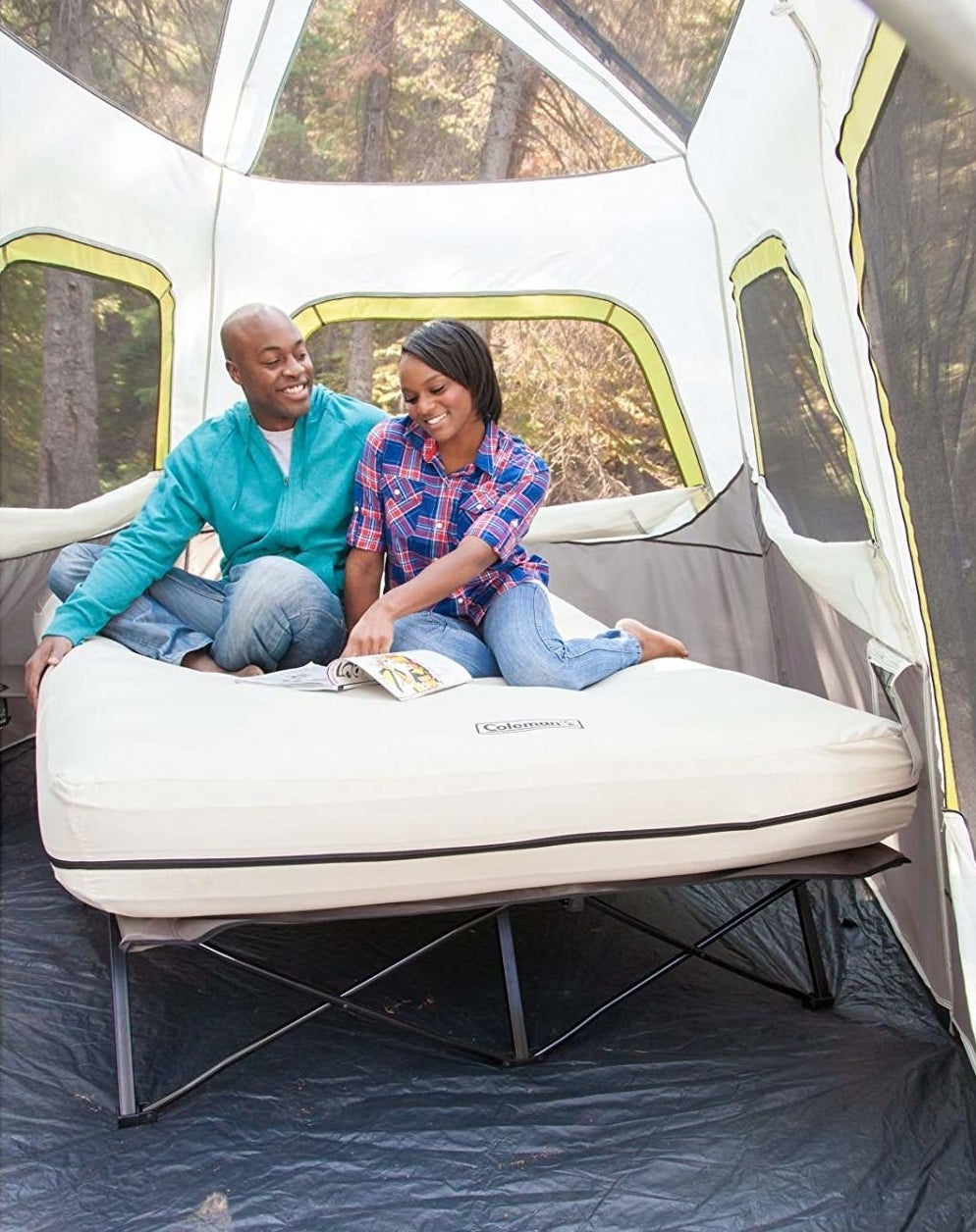 Two models sitting on a queen-size inflatable mattress on a cot inside a tall tent