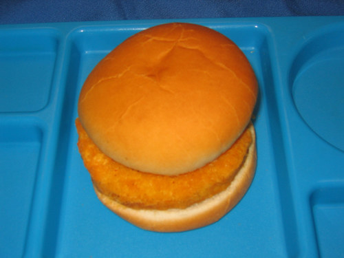 A fish patty sandwich in a tray with a blue background. The sandwich has a bread bun and a breaded fish patty