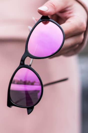 A hand holding black-framed sunglasses with mirrored pink lenses