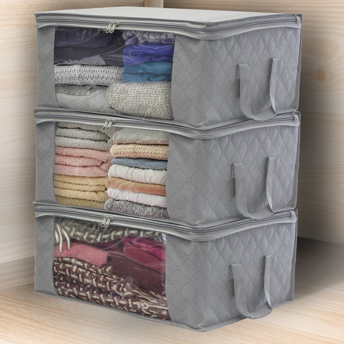 Three grey storage bags with clear panels on the side and handles on the front sitting on top of each other full of blankets and sweaters