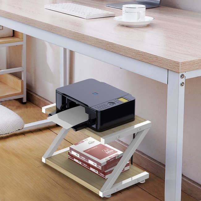 A two-tier shelf with a printer on the top shelf and a stack of paper on the bottom shelf under a desk