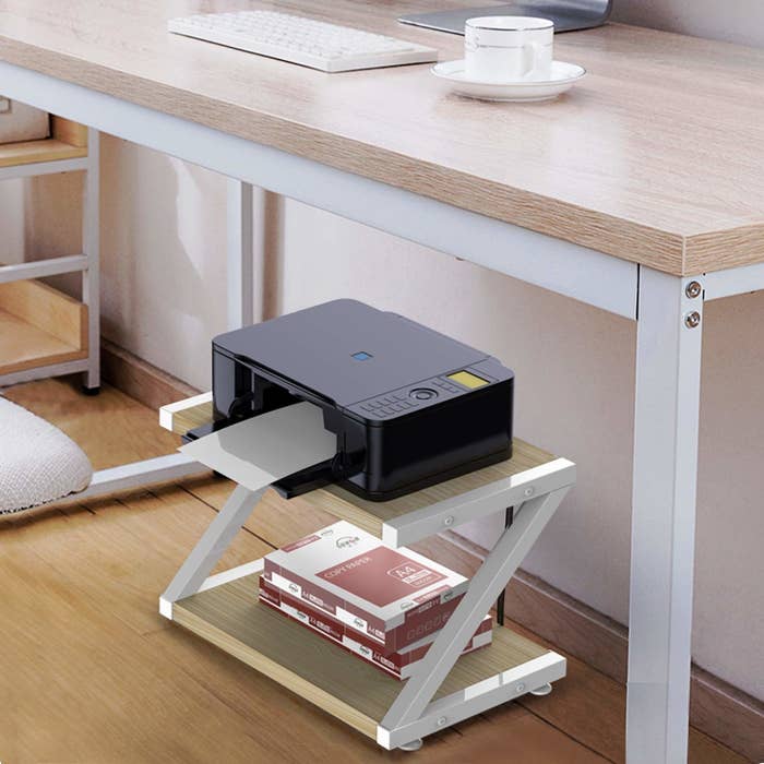 A two-tier shelf with a printer on the top shelf and a stack of paper on the bottom shelf under a desk