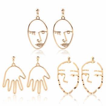 The three pairs of gold earrings: one pair that looks like abstract hands, one pair that looks like a face with no mouth, and one pair that looks like a winking face