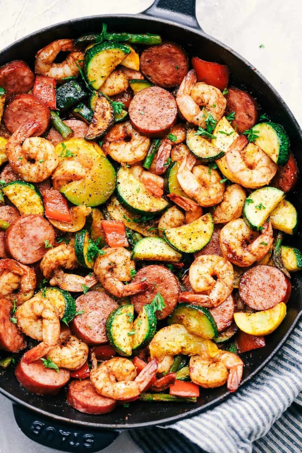 A skillet filled with a cooked mix of shrimp, sausage slices, zucchini, and bell peppers. The dish appears seasoned and garnished with herbs