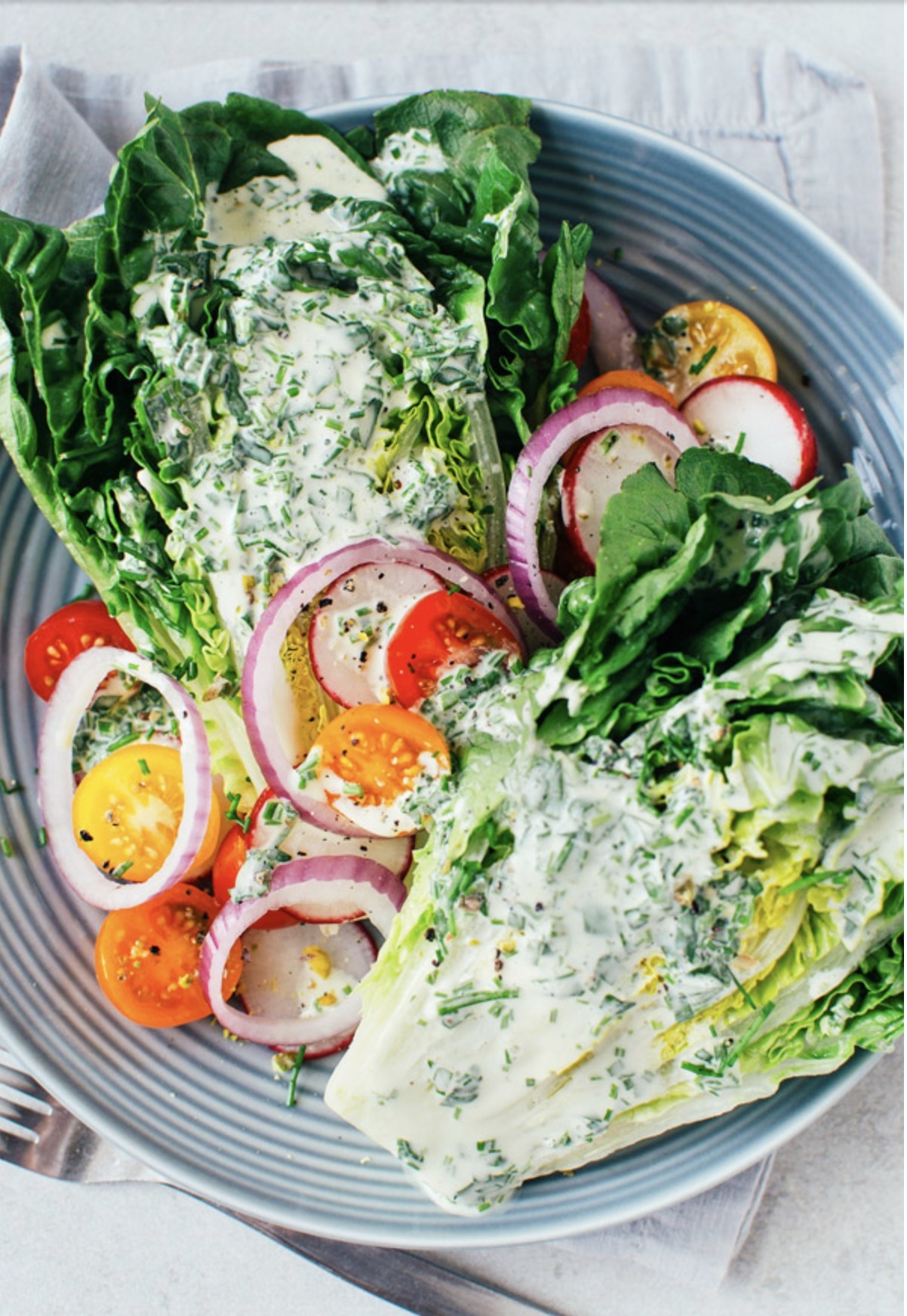 A plate of fresh salad with lettuce, sliced red onions, cherry tomatoes, and radishes, topped with a creamy herb dressing