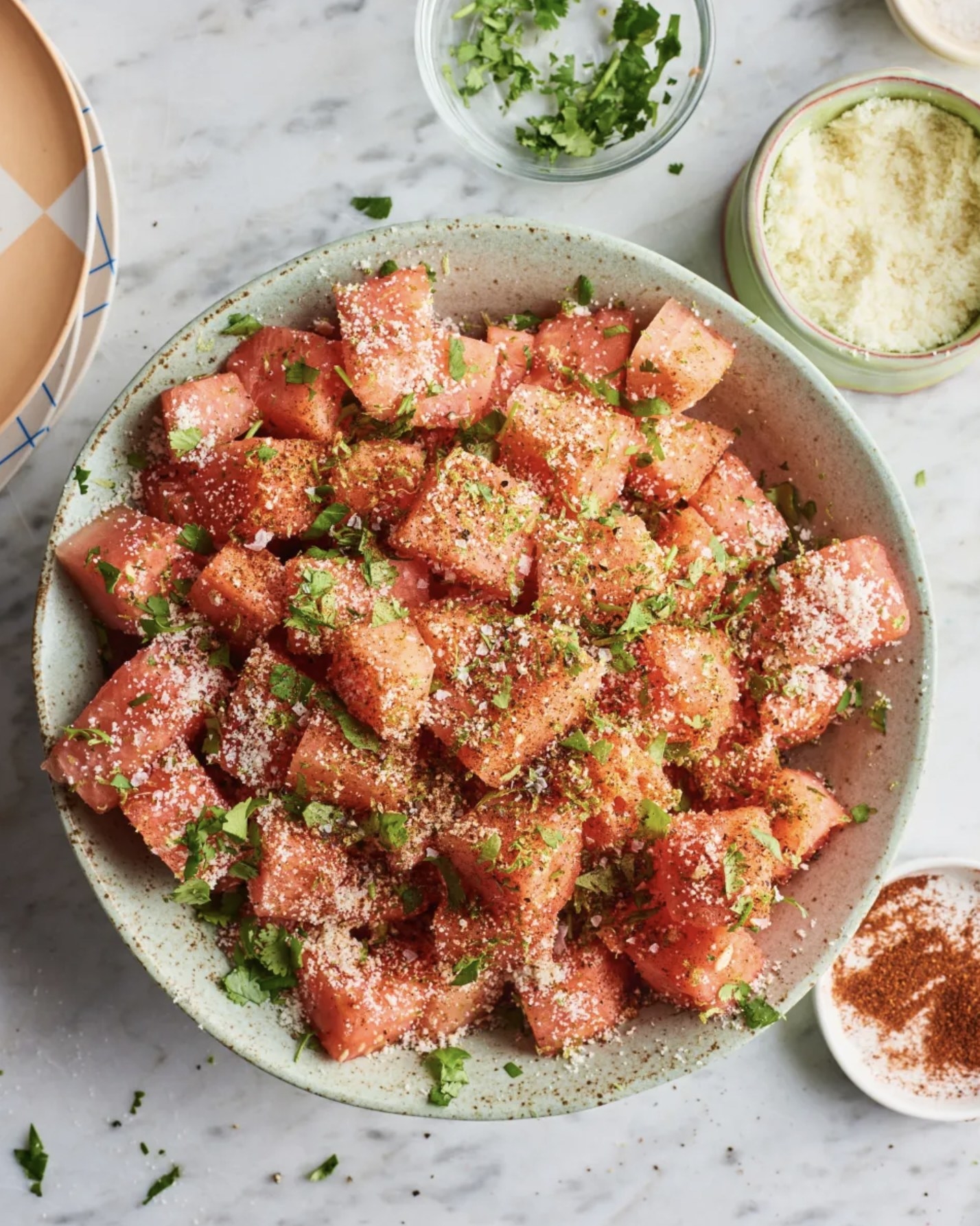 A bowl of watermelon cubes topped with chili powder, cheese, and fresh herbs, with additional garnishes in small bowls nearby