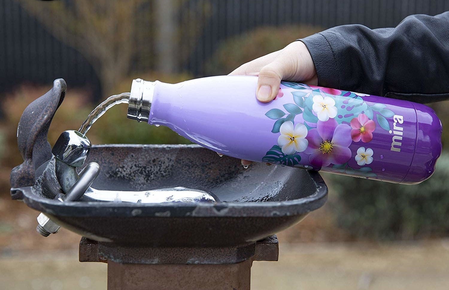 A hand refills a purple, narrow-necked stainless steel bottle (with purple, pink, and white flowers and green foliage print) at a water fountain