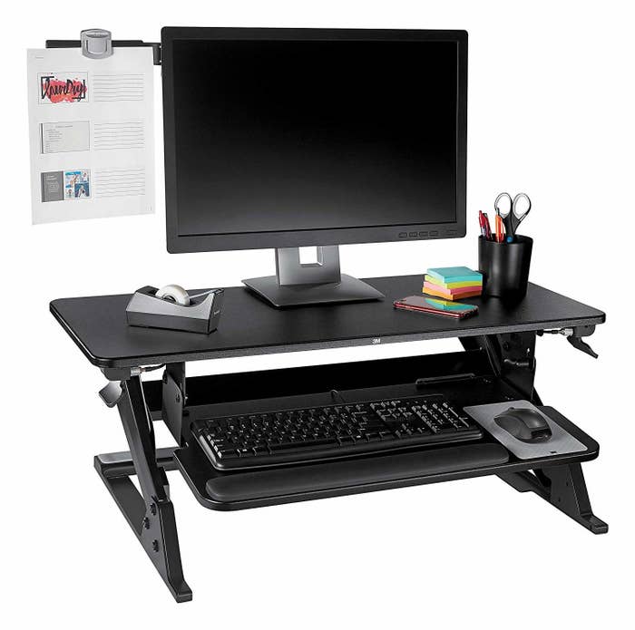 A monitor on a standing desk convertor with a clipboard-like arm extending from the side, holding a paper