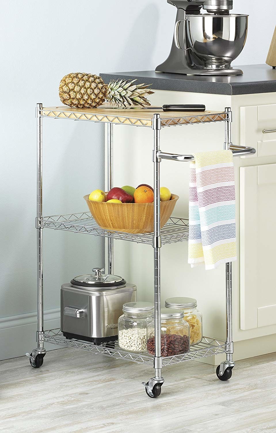 The silver metal three-tier kitchen cart with a wood cutting board on top and assorted kitchen products on it