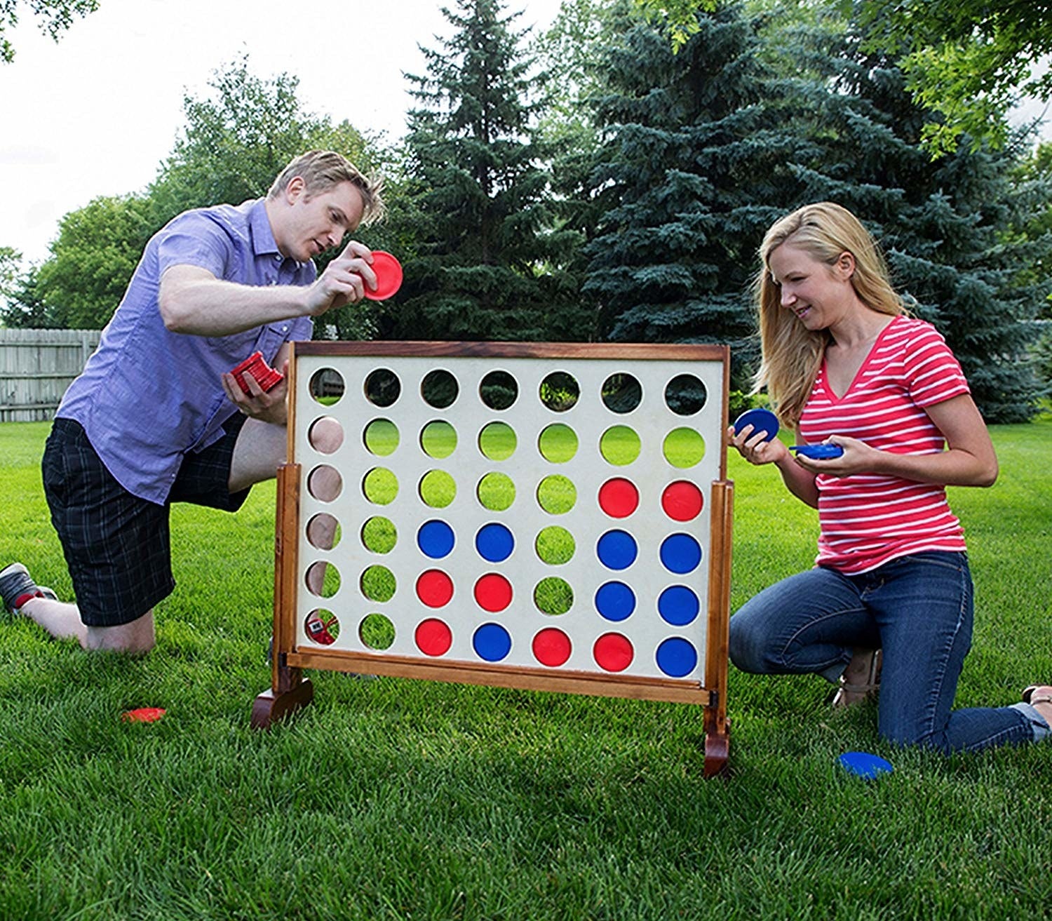a couple is kneeling in the grass playing with an oversized version of the game connect four. the board includes many open circle slots where blue and red tiles are place inside