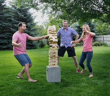 three people outside in the grass playing with a very tall tower of blocks