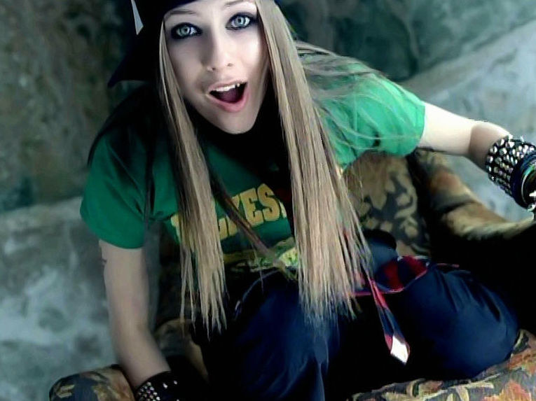 Avril sits on her knees on a couch wearing a tie and lots of bracelets