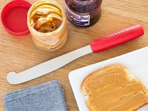 The knife with a curved end and red handle sitting on a table with a jar of peanut butter and jelly and a piece of peanut butter toast